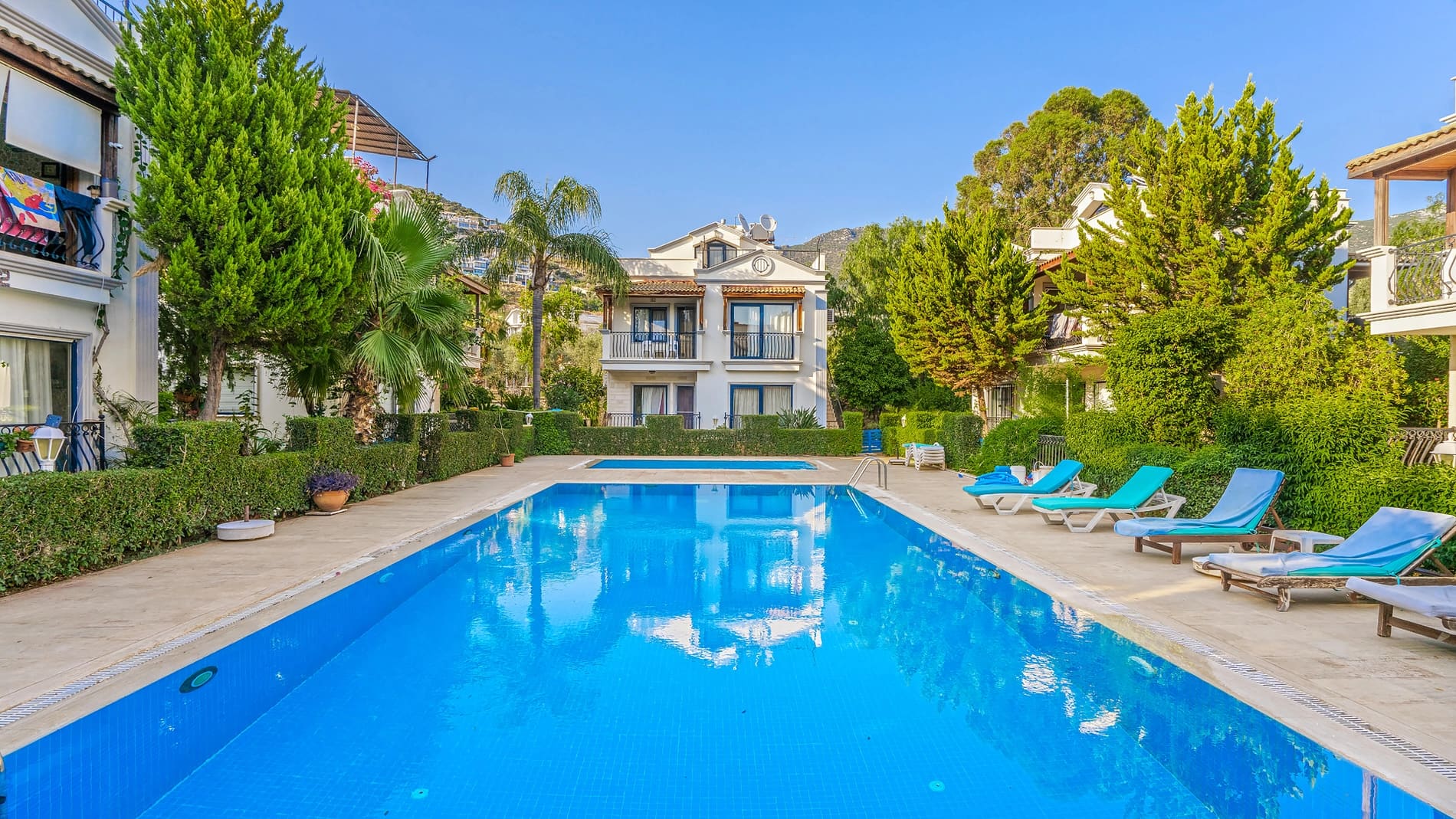Ağaçlar ve şezlonglarla çevrili açık havuz ve arka planda villa