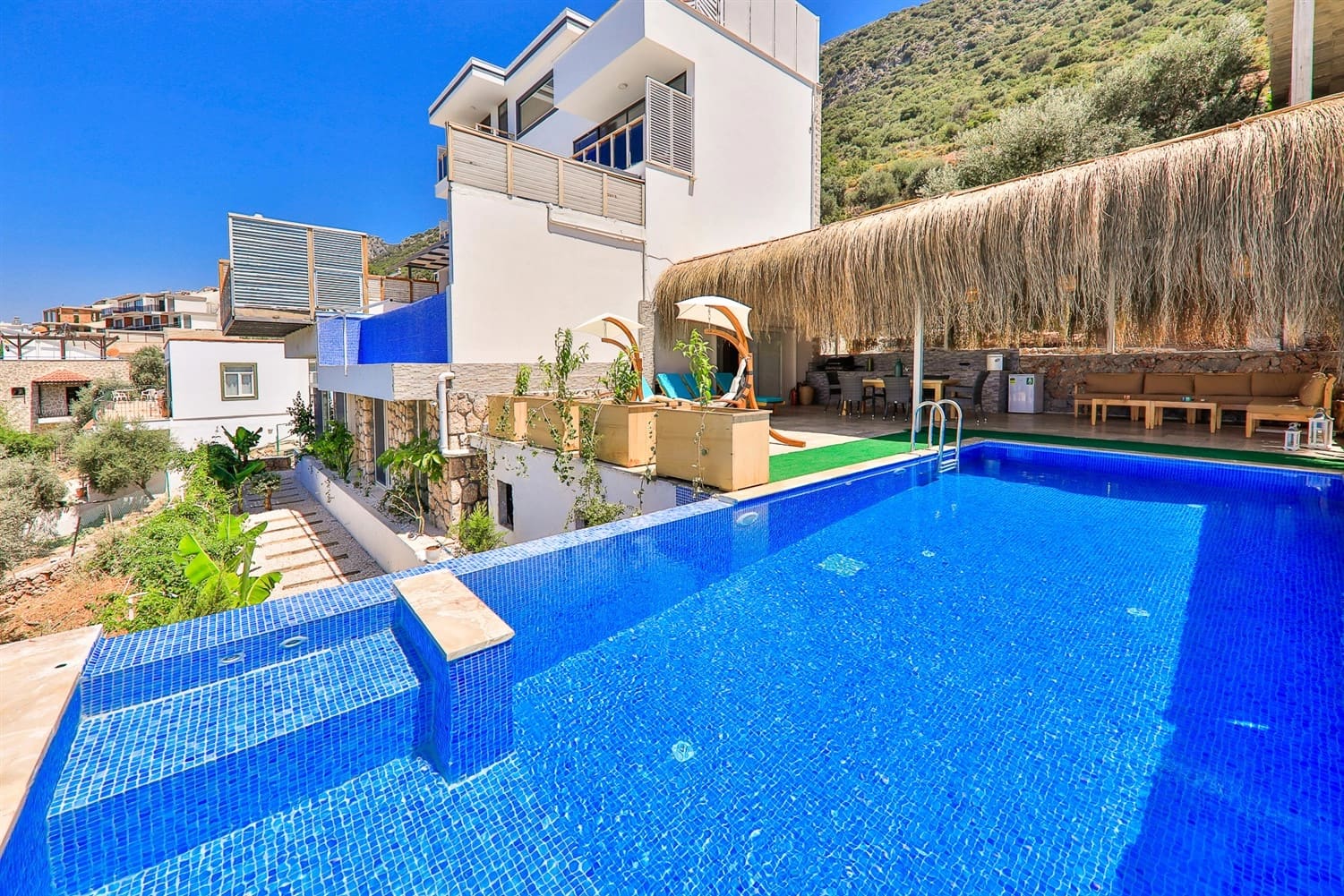 Hasır gölgelikli oturma alanı ve havuz başında şezlongların bulunduğu, deniz manzaralı villa.