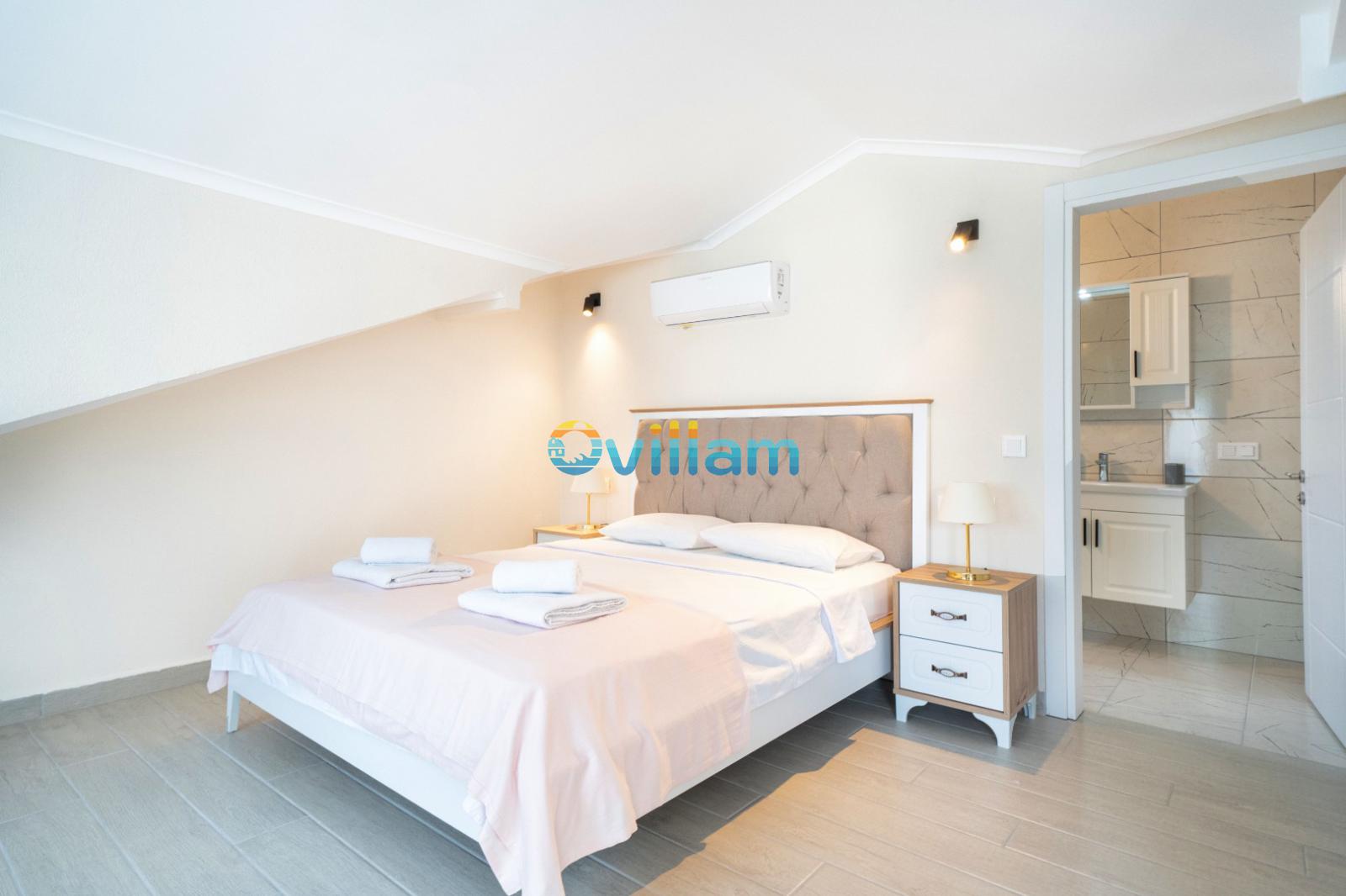 Çift kişilik yatak, komodinler ve yanında banyo kapısı bulunan aydınlık yatak odası.