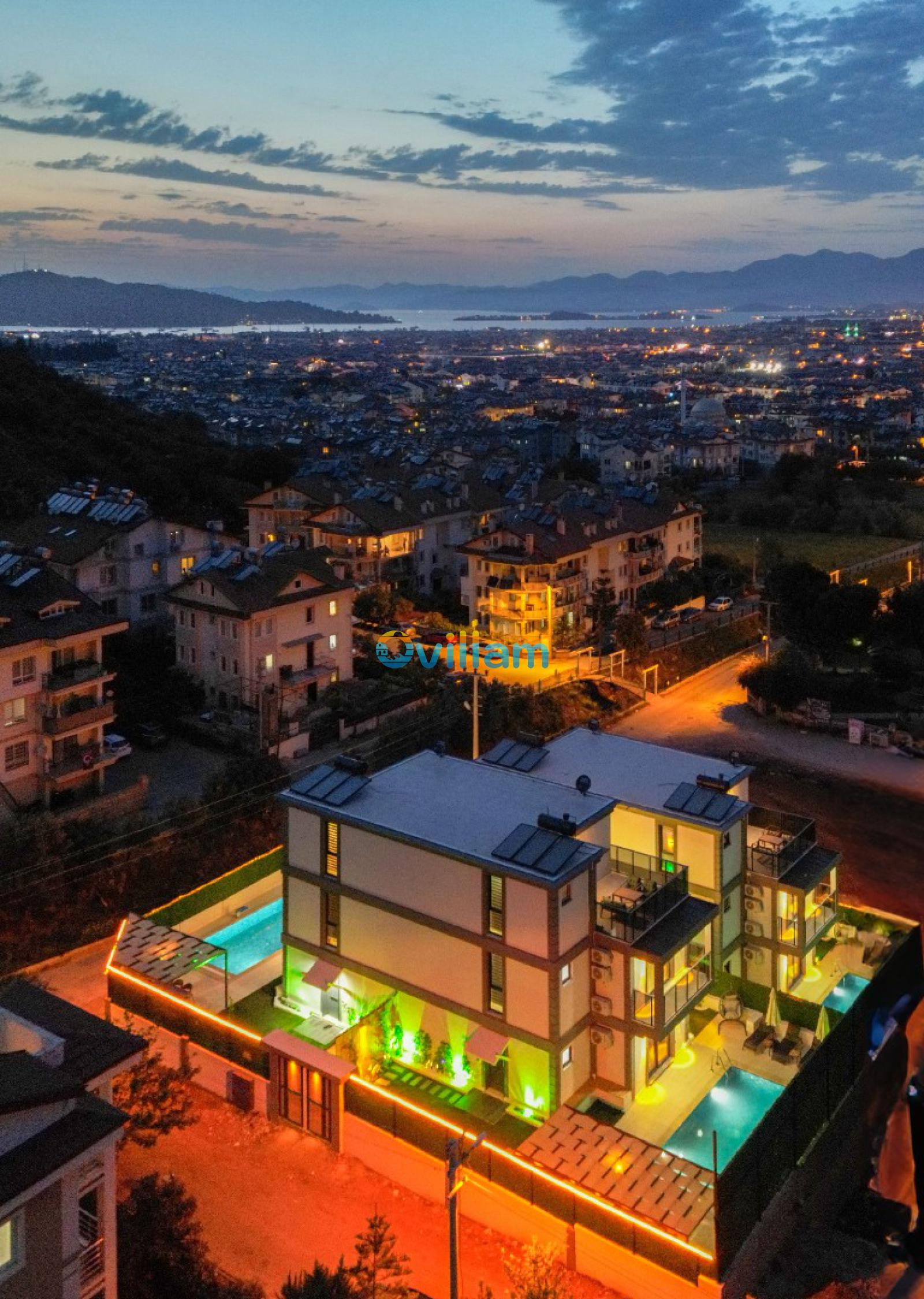 Gece ışıklandırılmış villa, havuzlar ve arkada şehir ile deniz manzarası.