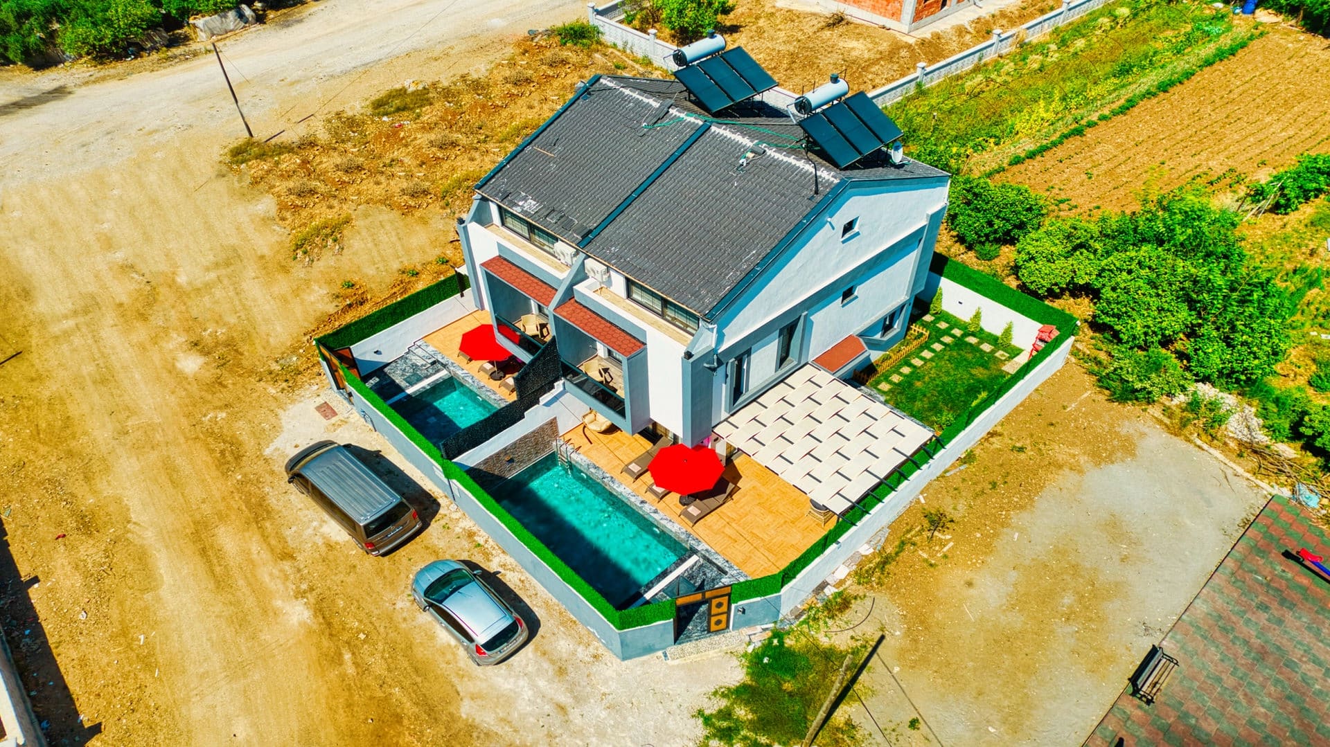 İki bitişik villanın havuzlu bahçeleri, güneş panelleri ve park halindeki araçların kuş bakışı görünümü.