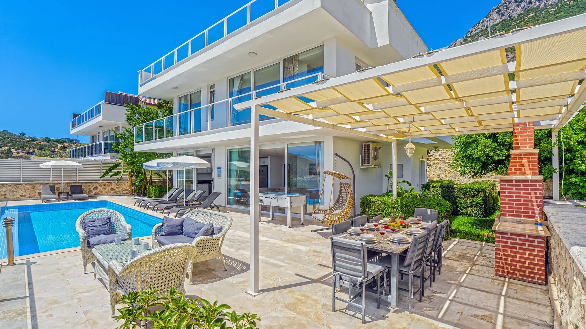 Havuz kenarında şezlonglar, yemek masası, barbekü ve gölgelik bulunan modern villanın dış görünümü.