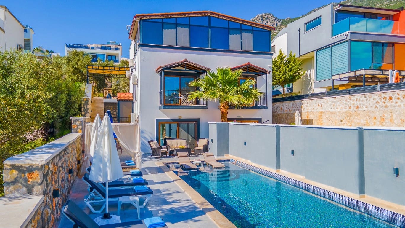 Taş duvarlı terasın önünde uzun bir havuz, şezlonglar ve arkasında beyaz dış cepheli villa.