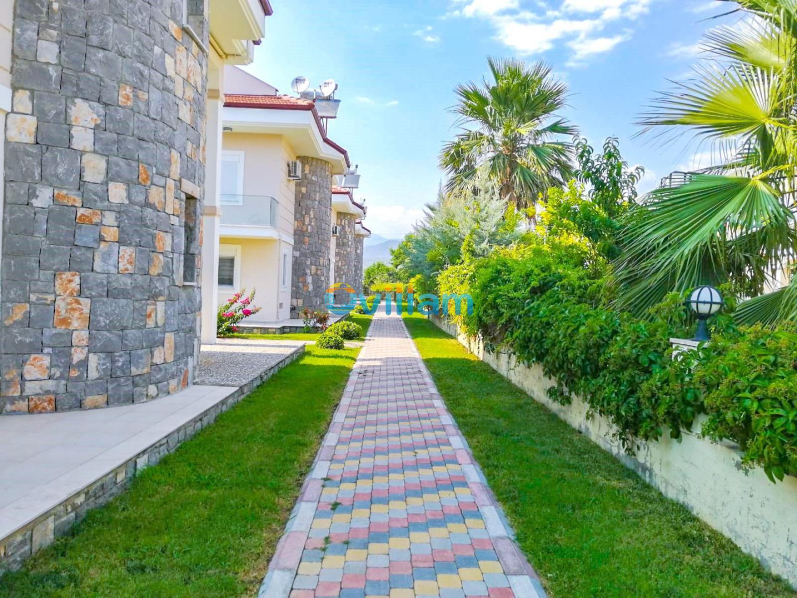 Taş kaplamalı villa cepheleri arasında uzanan renkli parke taşlı yürüyüş yolu ve yanındaki yeşil alan.