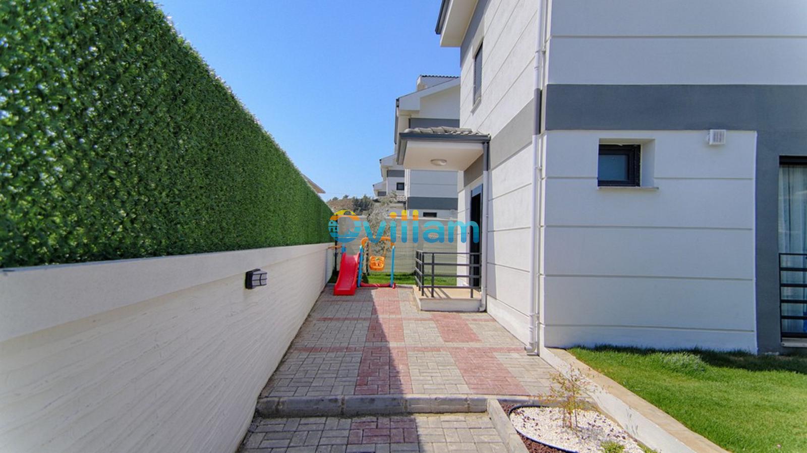 Villa bahçesinde yeşil çitlerle çevrili, çocuk kaydırağı bulunan taş döşeli yan bahçe alanı.