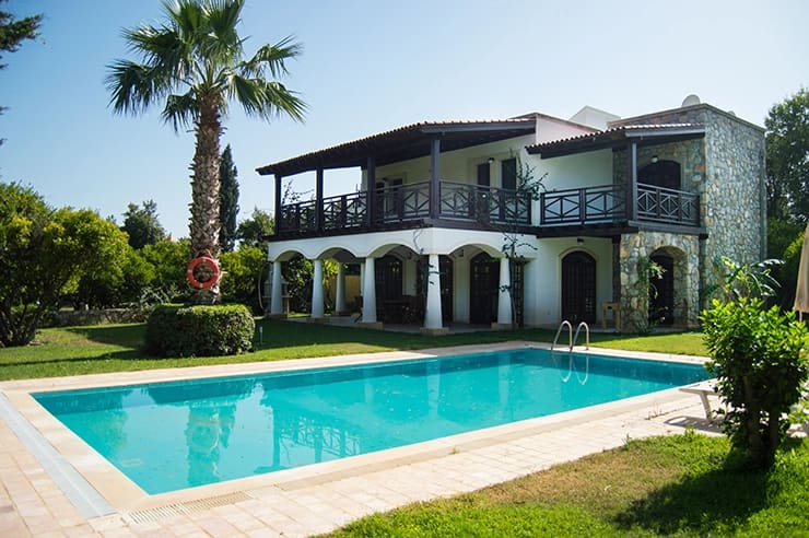 Bahçesinde havuz ve palmiye ağacı bulunan iki katlı taş villa dış cephesi.