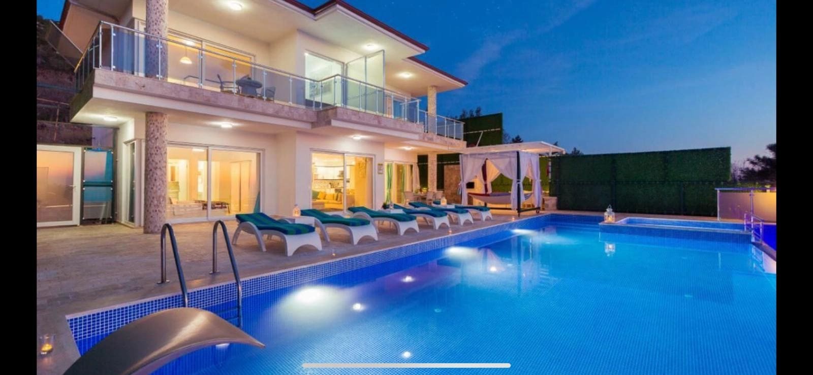 Aydınlatmalı havuz kenarında şezlonglar bulunan iki katlı villa dış cephesi.