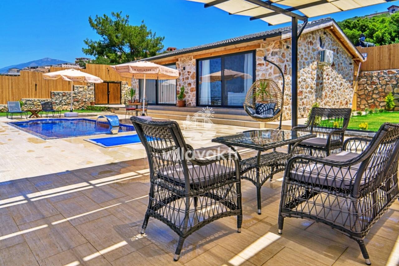 Taş villa önünde havuz, rattan oturma grubu, salıncak ve güneş şemsiyeleri bulunan geniş bir teras.