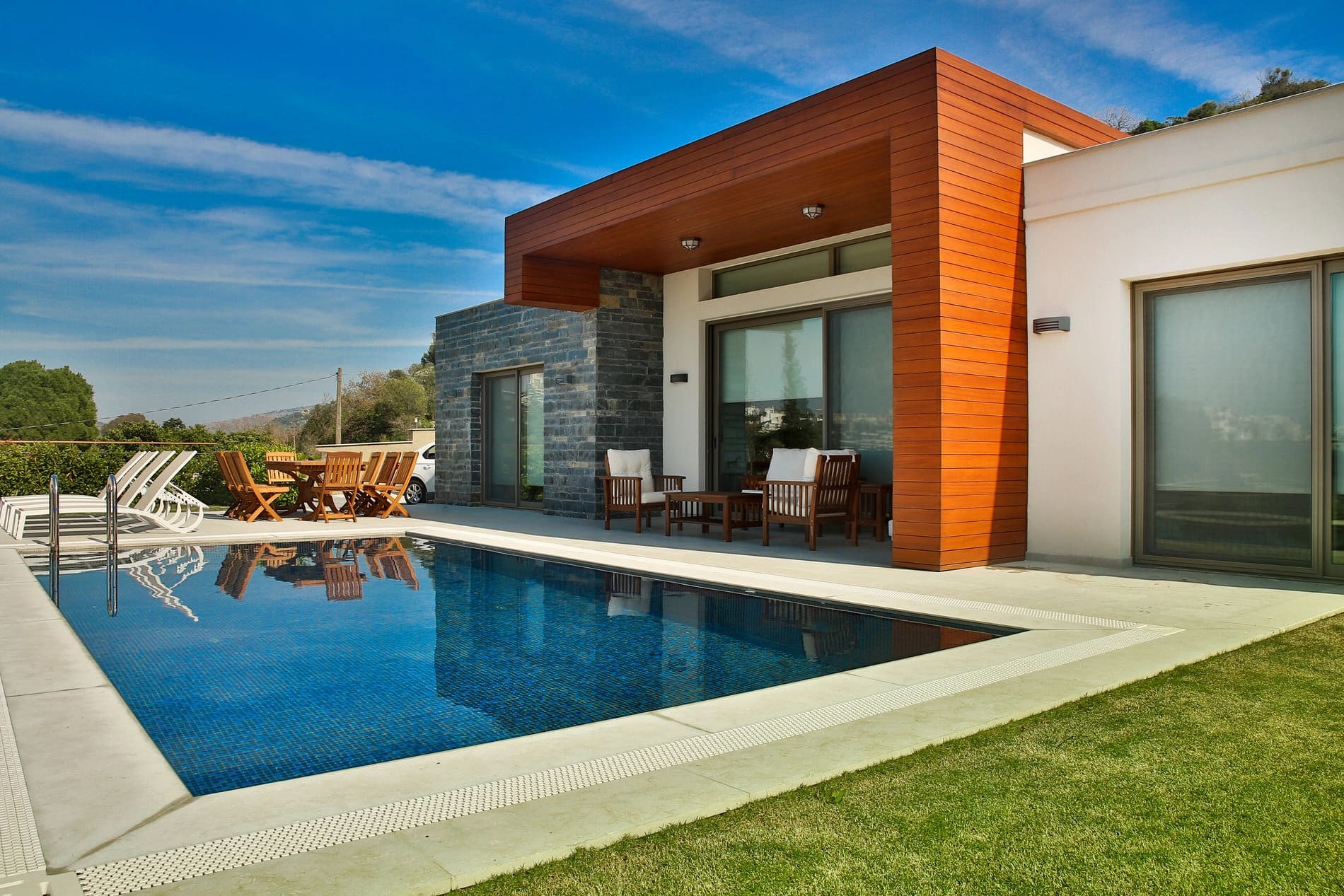 Ahşap detaylı modern villa cephesi, önünde mavi yüzme havuzu, şezlonglar ve yemek masası bulunuyor.