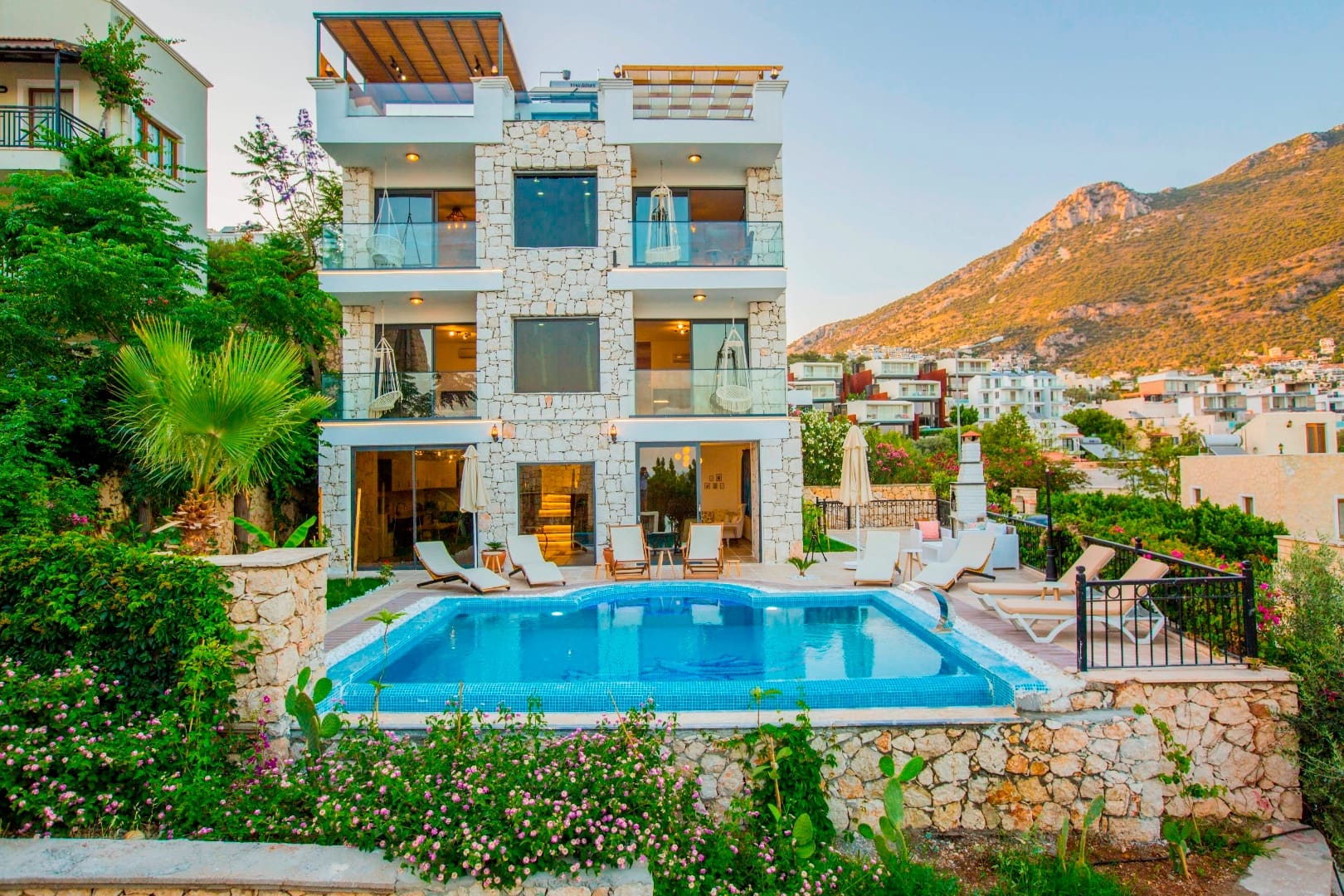 Taş kaplamalı, üç katlı villa, önünde yüzme havuzu, şezlonglar ve dağ manzarası.