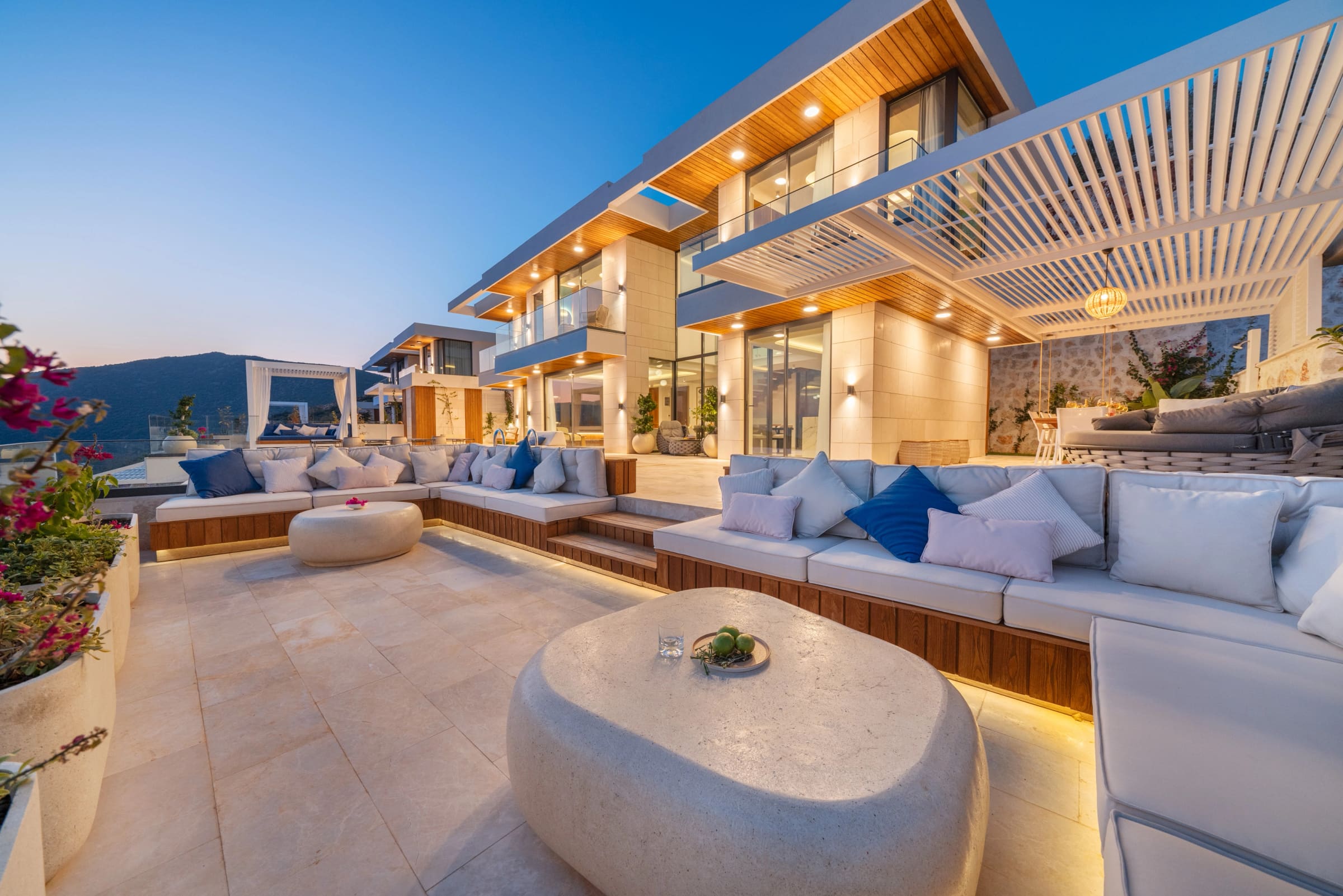 Akşam ışıklandırmalı, geniş oturma grupları ve taş masalarla döşenmiş modern villa terası.
