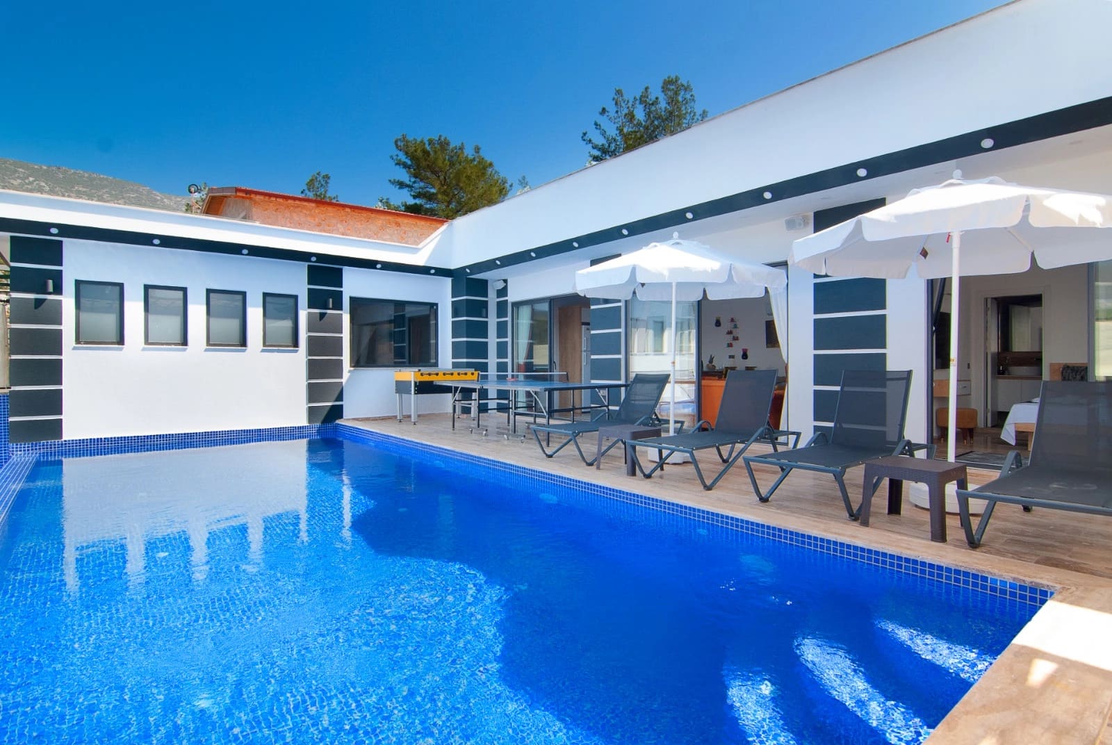 Mavi fayanslı özel havuz, şezlonglar, güneş şemsiyeleri ve masa tenisi bulunan villa terası.