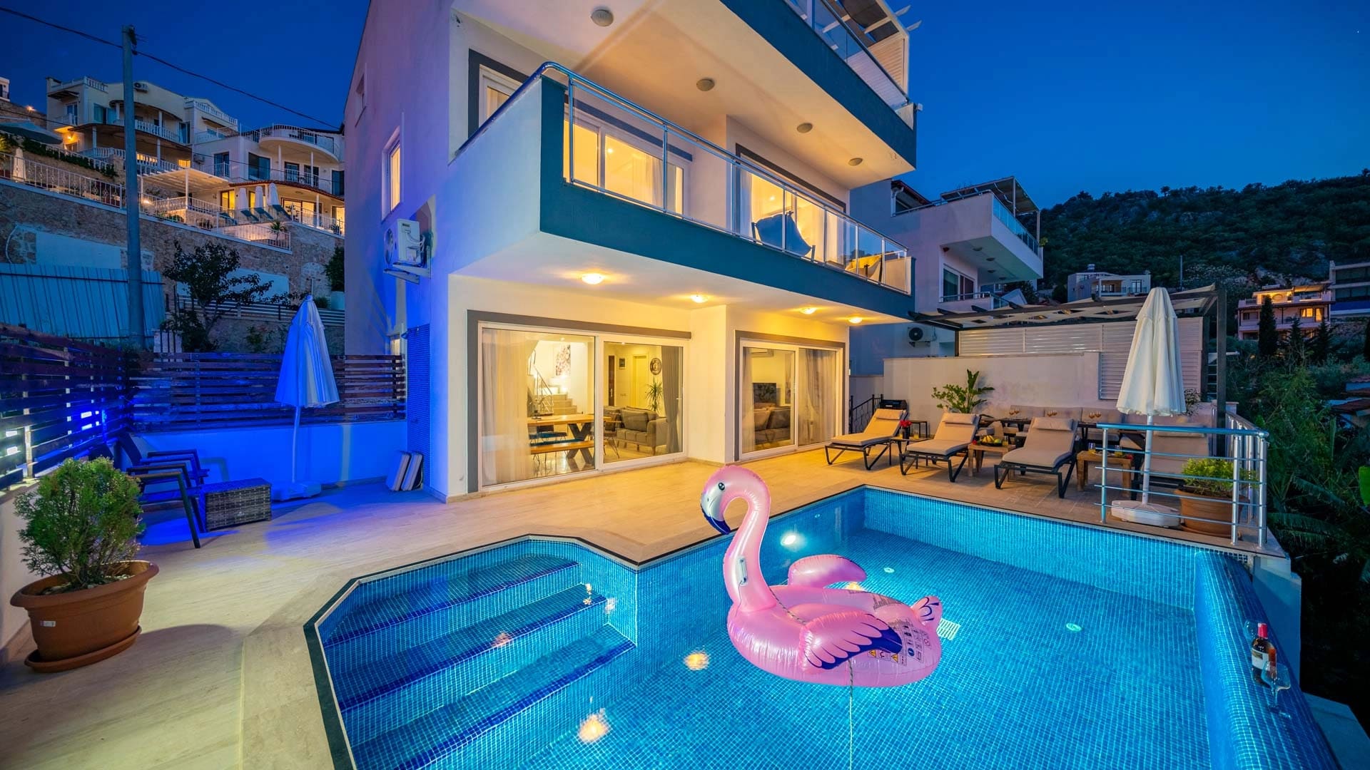 Akşam ışıklandırmalı havuz başında pembe flamingo simit ve şezlonglar.