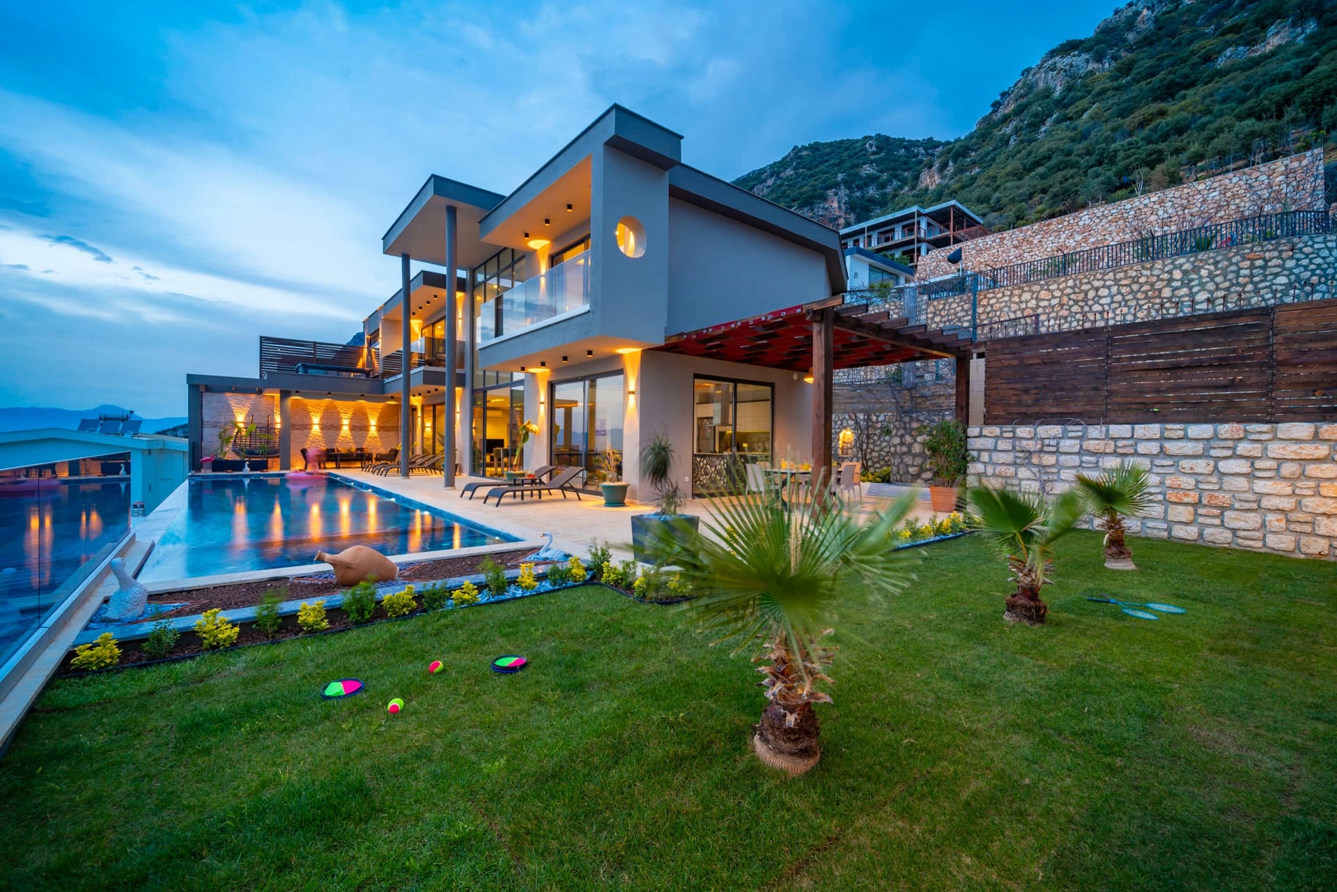 Akşam ışıklandırmalı modern villa, sonsuzluk havuzu, yeşil bahçe ve taş duvarlı dış cephe.