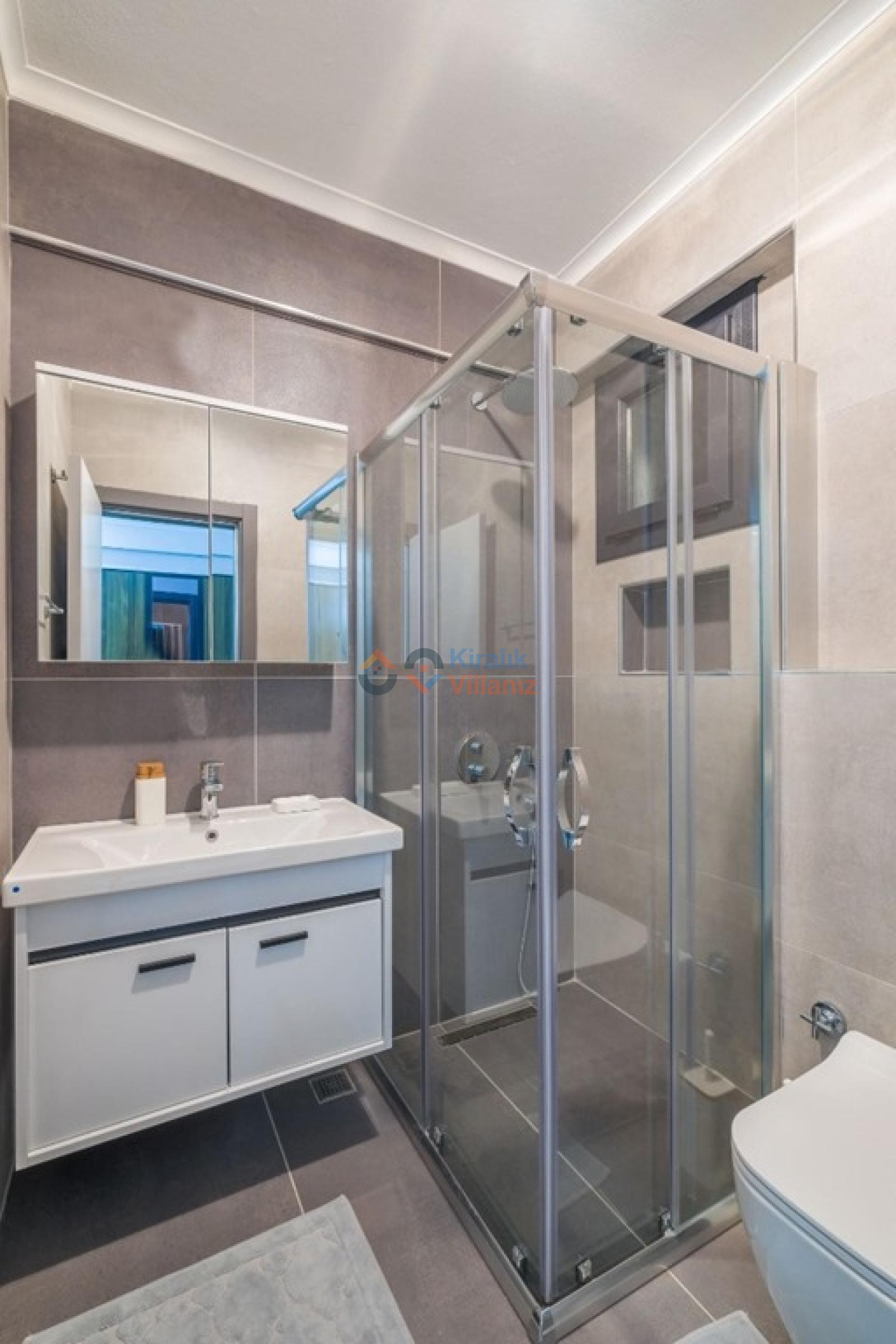 Gri fayanslarla kaplı banyoda cam duşakabin, beyaz dolaplı lavabo ve aynalı dolap bulunuyor.