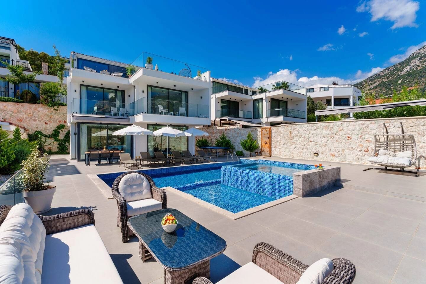 Modern villanın önünde basamaklı havuz, şezlonglar, oturma grubu ve güneş şemsiyeleri bulunuyor.