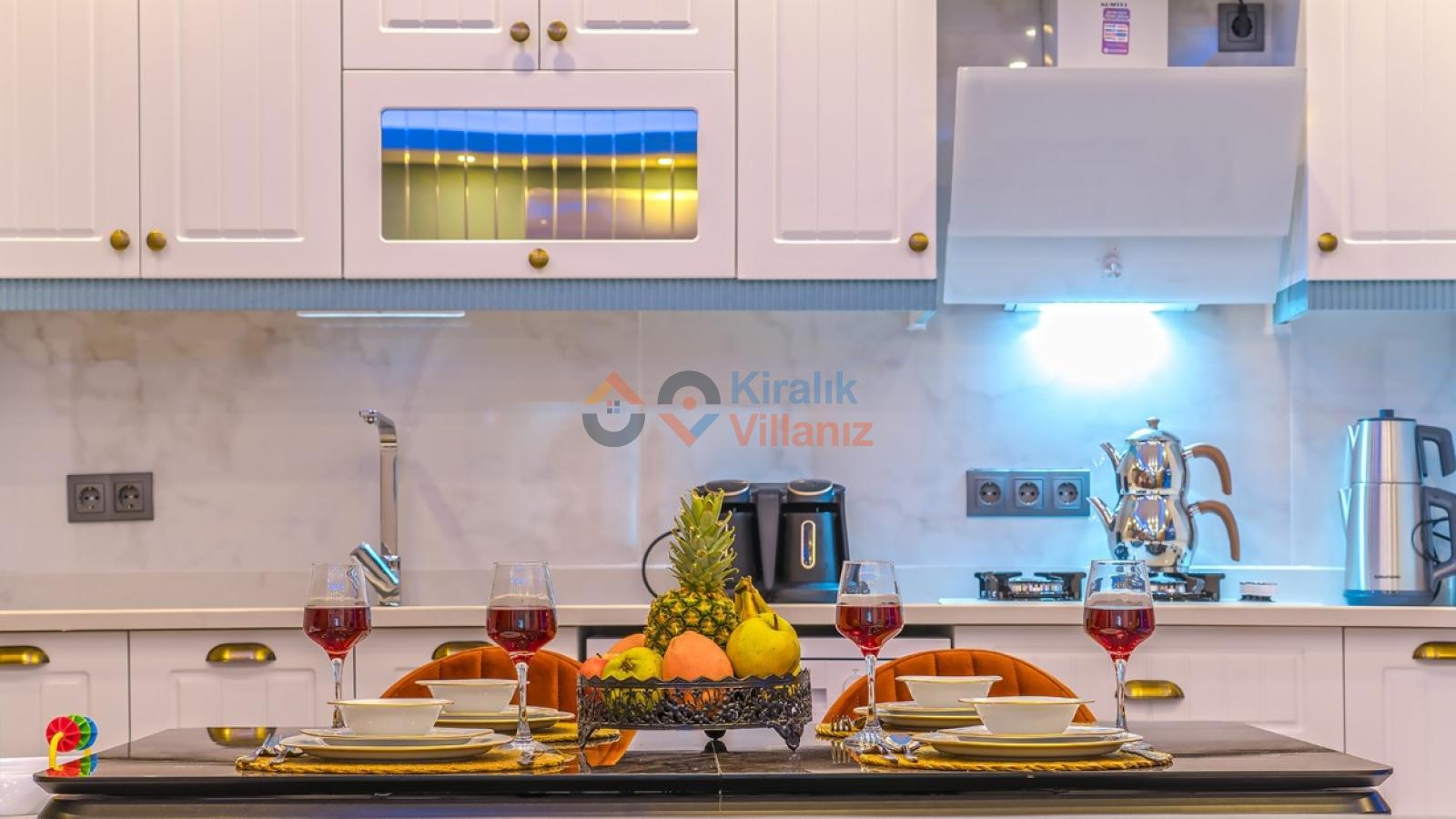 Beyaz mutfak dolapları, üzerinde meyve tabağı ve kadehler bulunan şık bir yemek masası.