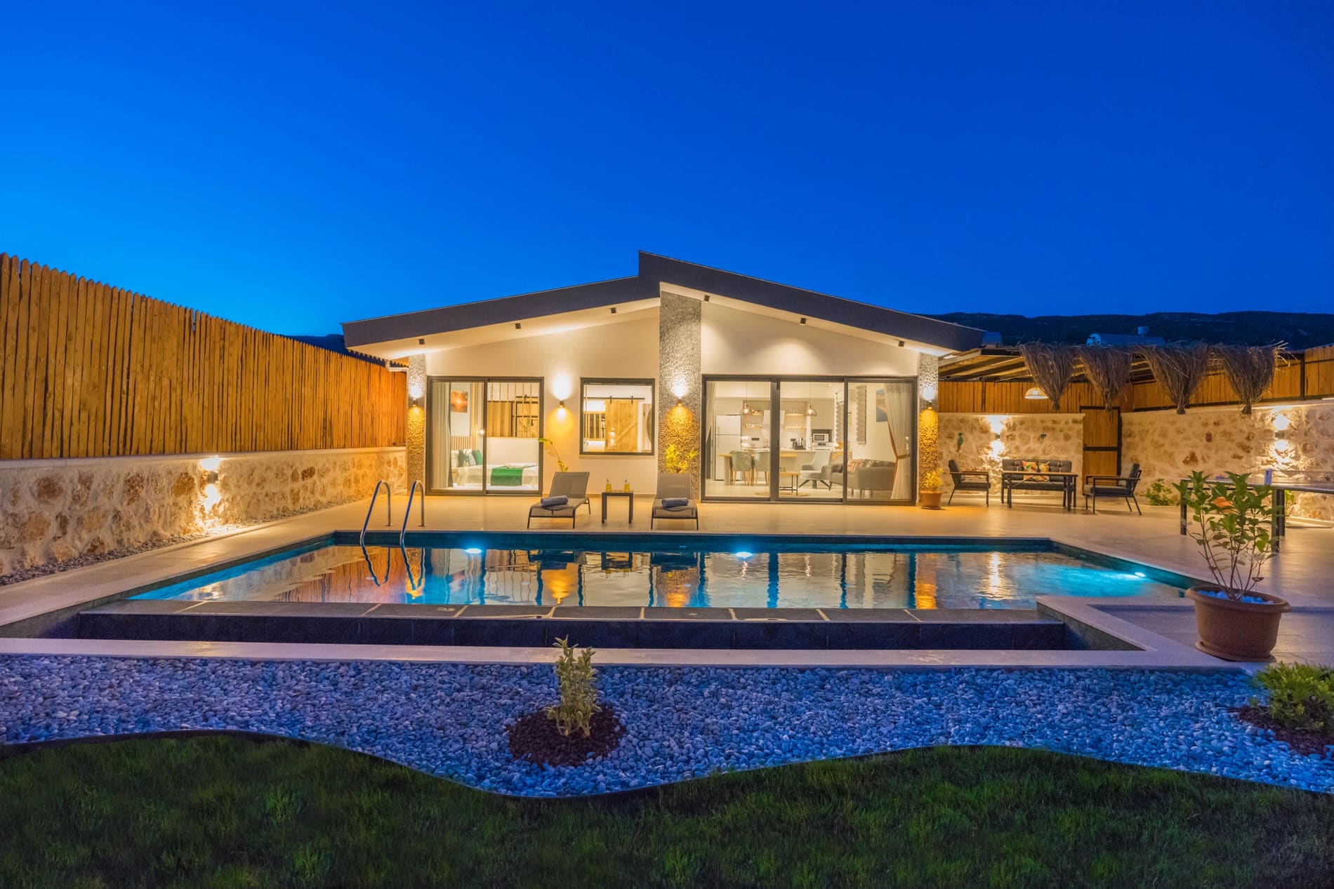 Gece aydınlatılmış modern villa, önünde dikdörtgen havuz ve bahçe mobilyaları.