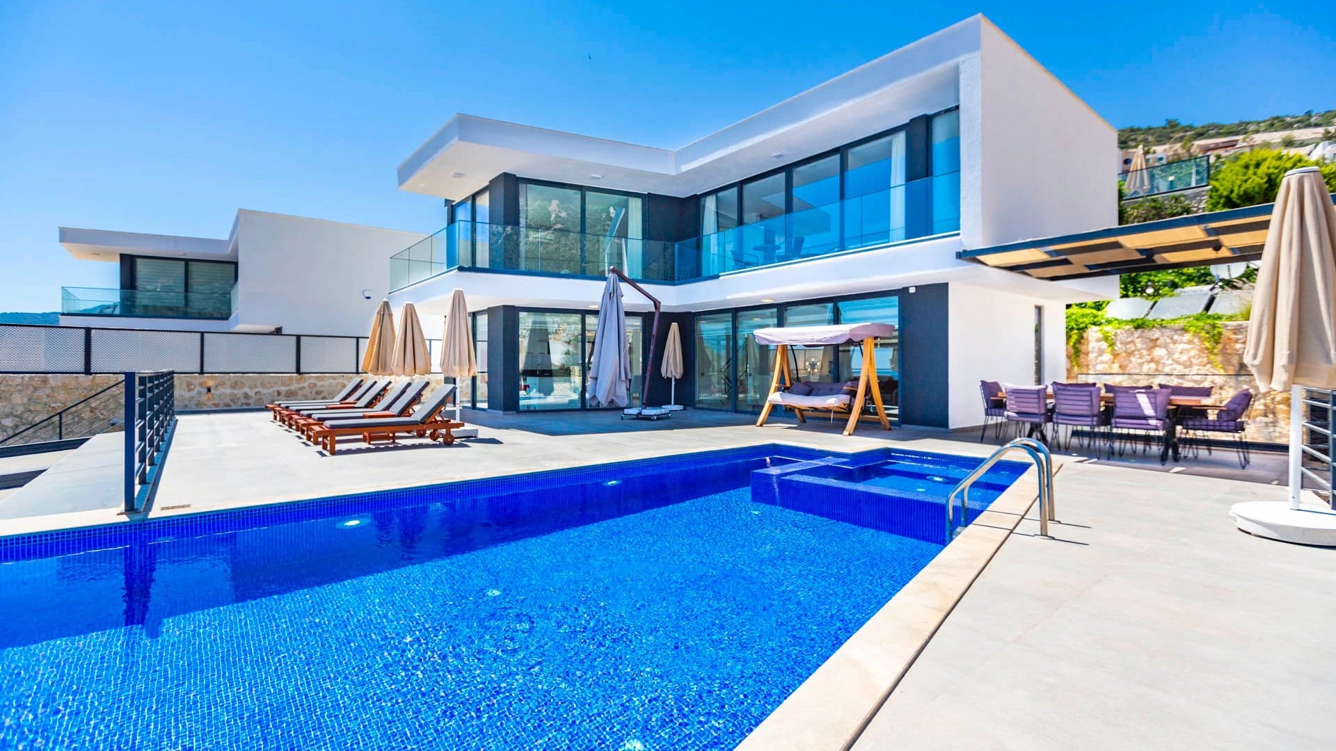 Modern beyaz villa, önünde mavi havuz, şezlonglar, salıncak ve yemek masası bulunan geniş teras.