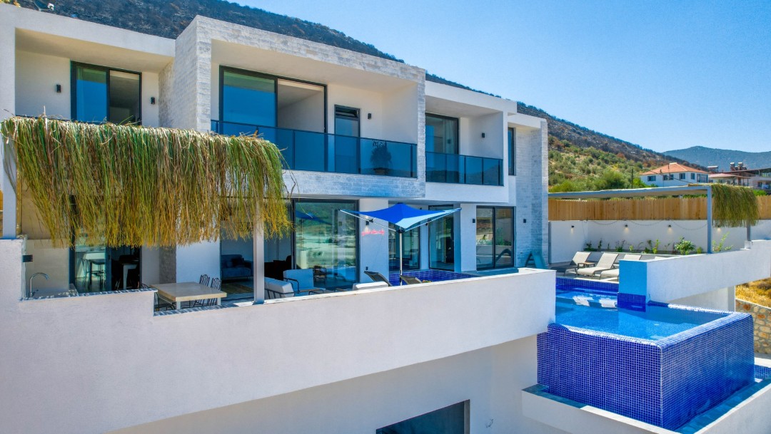 Aydınlık havada dağ yamacında yer alan, balkonlu ve mavi havuzlu beyaz dış cepheli iki katlı modern villa.