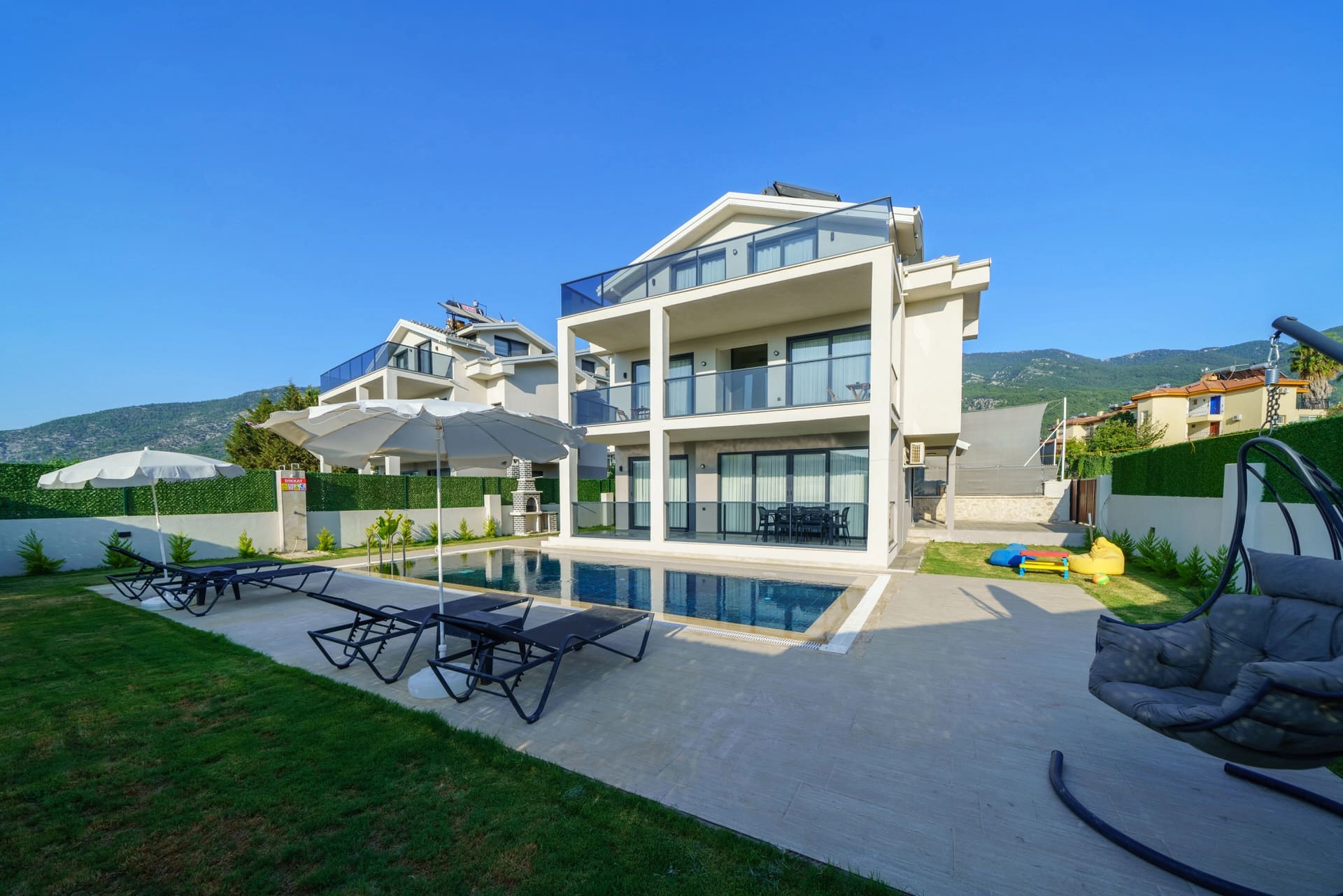 Modern beyaz villanın önünde yüzme havuzu, şezlonglar, güneş şemsiyeleri ve bahçe alanı.