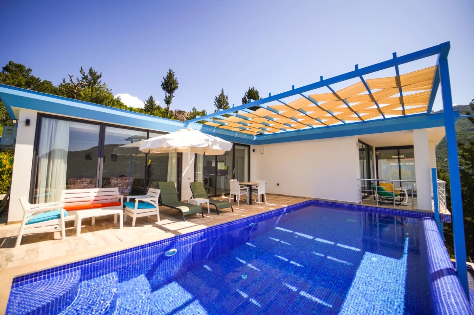 Mavi gölgelikli terası, yüzme havuzu ve dış mekan oturma grubu olan beyaz villa.