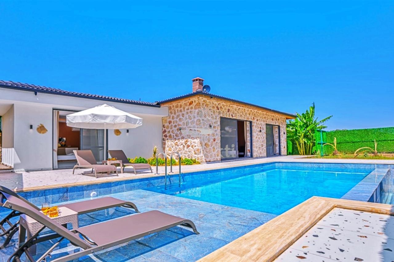 Taş duvarlı villa, önünde mavi yüzme havuzu, şezlonglar ve güneş şemsiyesi bulunan geniş bir teras.
