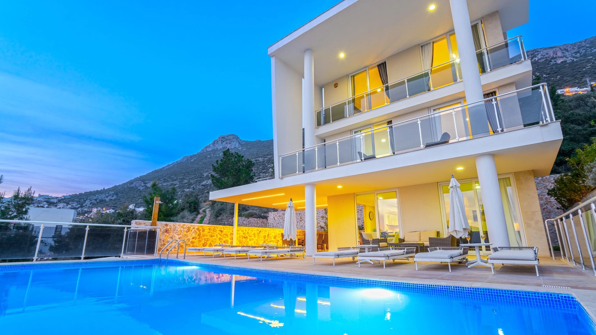 Akşam ışıklandırmalı, üç katlı modern villanın önünde mavi havuz ve şezlonglar.