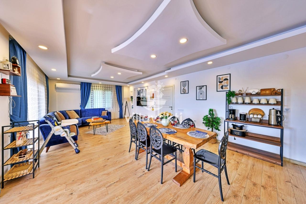 Modern salon ve yemek odasında ahşap yemek masası, mavi koltuk takımı ve açık mutfak rafı bulunuyor.