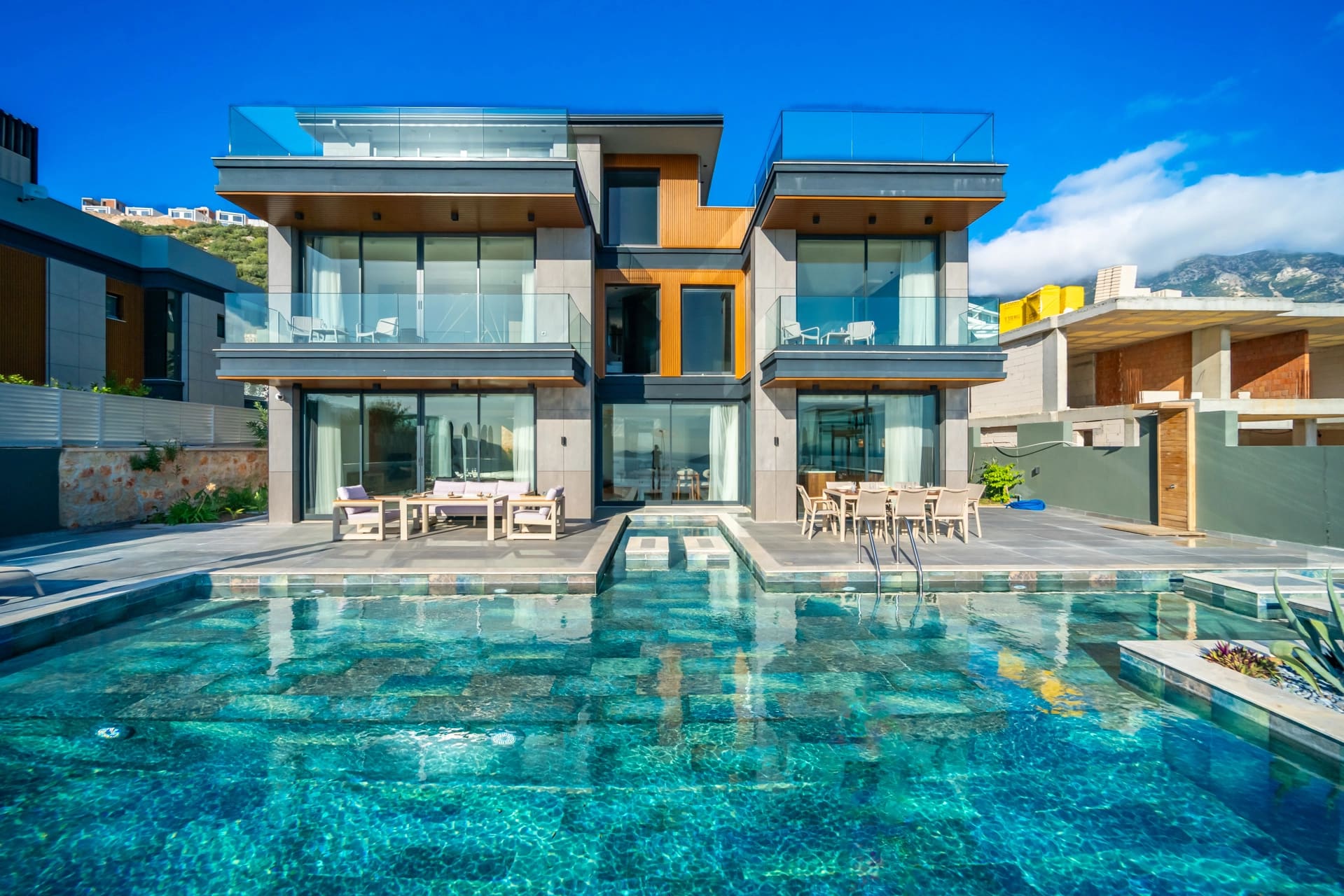 Modern mimariye sahip villa, önünde basamaklı havuz ve güneşlenme terasında yemek masası bulunuyor.