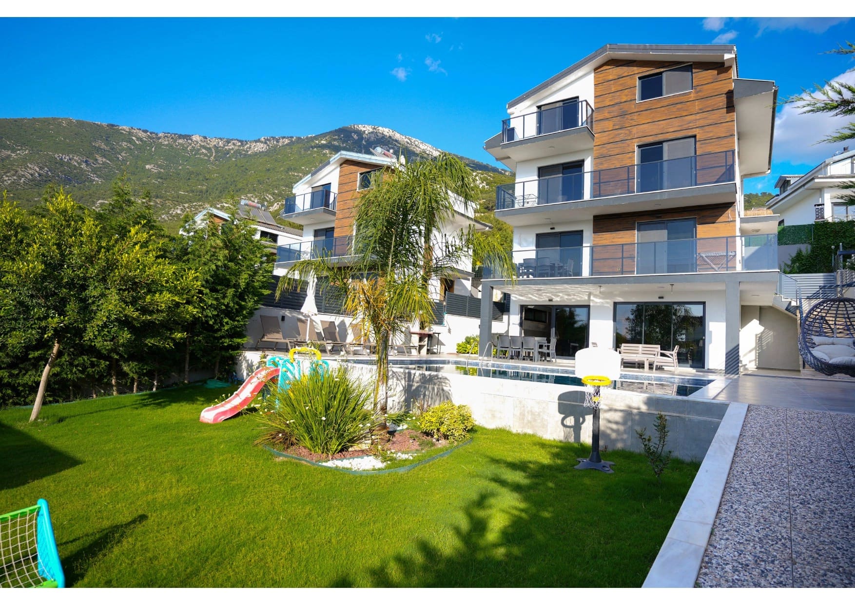 Üç katlı modern villa, bahçesinde çocuk oyun alanı, havuz ve dağ manzarası ile dikkat çekiyor.