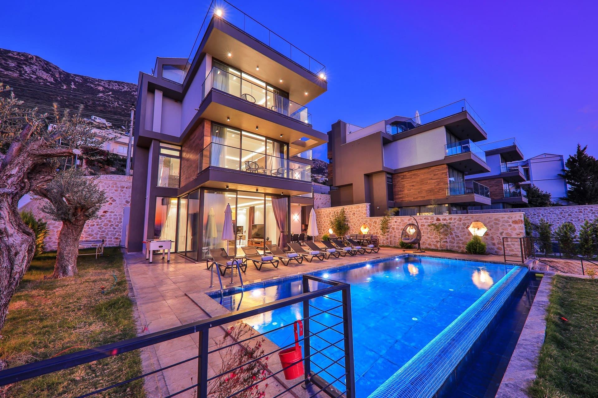 Akşam ışıklandırmalı modern villa, önünde ışıklı yüzme havuzu ve şezlonglar.
