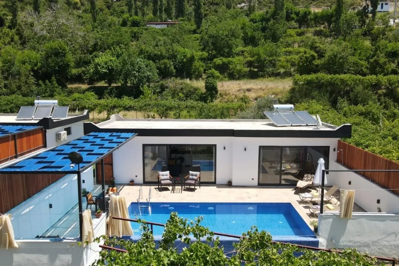 Ağaçlar arasında havuzlu ve şezlonglu bahçesi olan tek katlı beyaz villa.