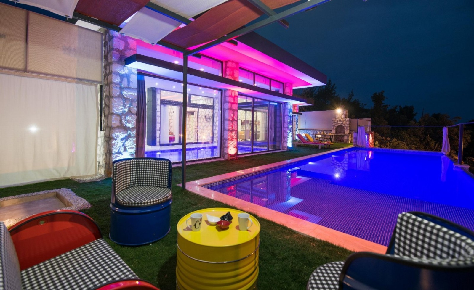 Gece pembe ve mavi ışıklandırmalı havuz kenarında oturma grubu ve villa dış cephesi.
