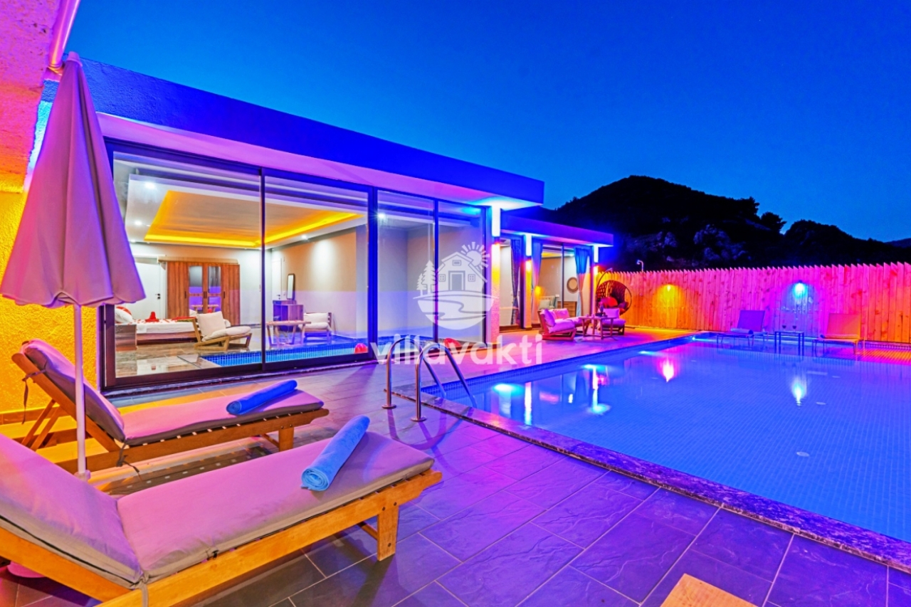 Gece aydınlatmalı özel havuz, ahşap şezlonglar ve cam kapılı modern villa dış cephesi.
