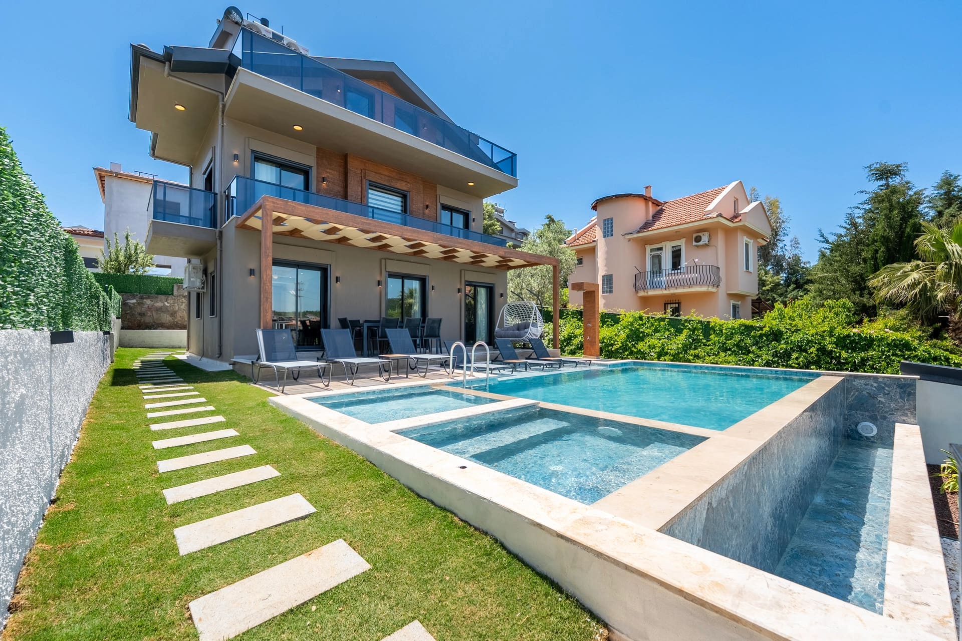 Modern villa, özel havuz, şezlonglar ve bahçe yolu ile dış cephe görünümü.