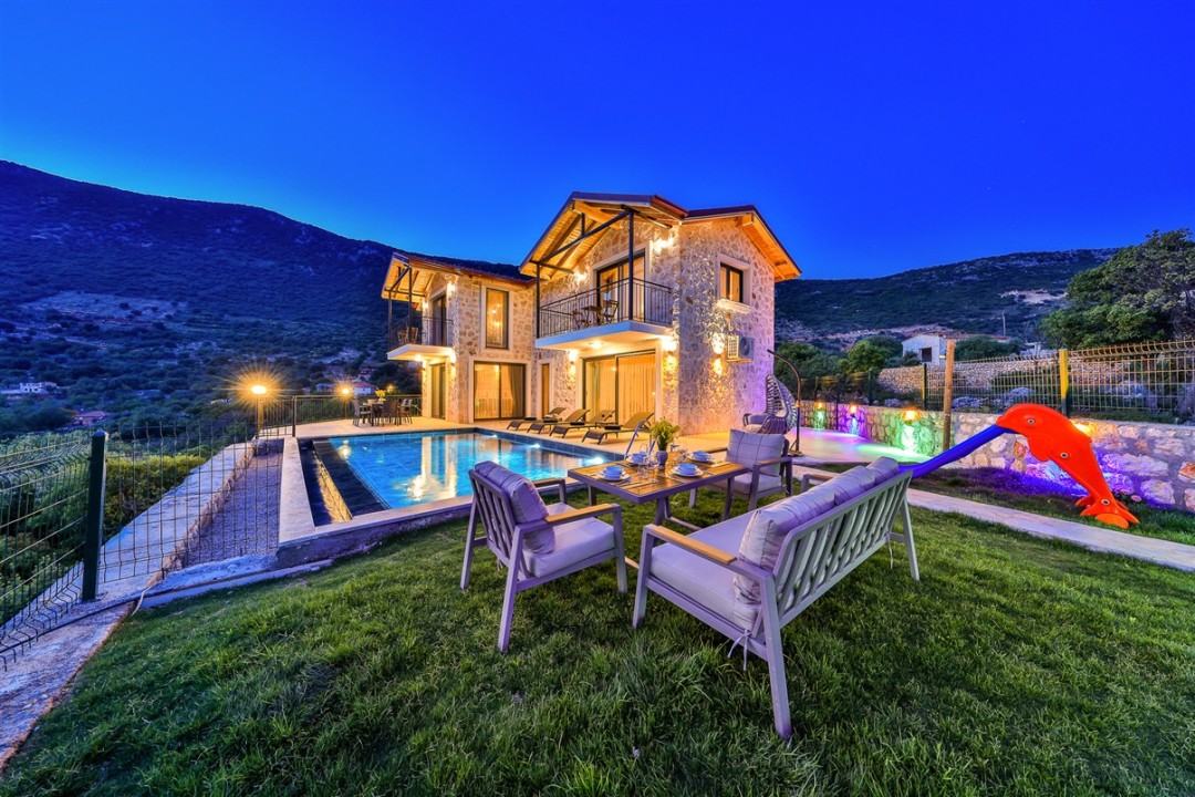 Akşam vakti aydınlatılmış taş villa, havuz, bahçe oturma grubu ve dağ manzarası.