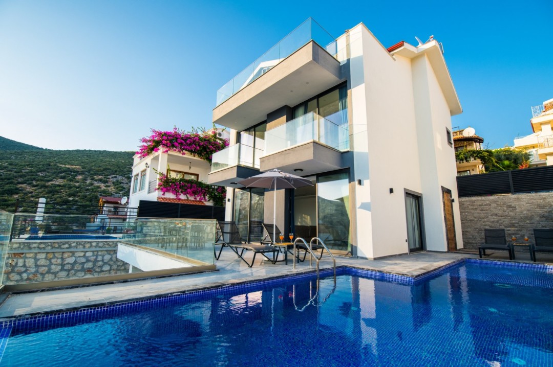 Mavi havuzlu, iki katlı modern villanın önünde şezlonglar ve şemsiye var.