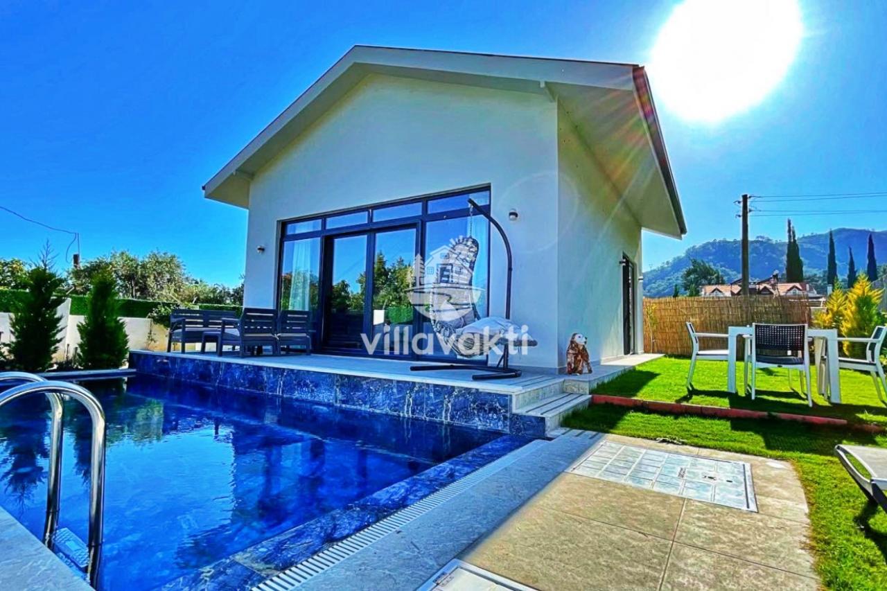 Mavi havuzlu, geniş cam kapılı, bahçeli ve güneşli bir villa dış cephesi.