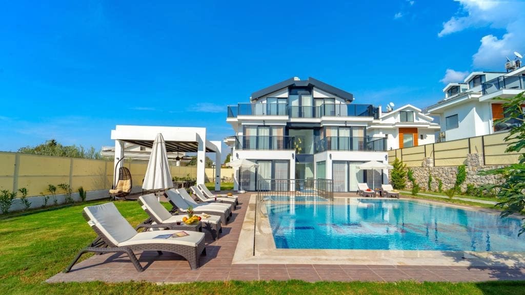Bahçesinde şezlonglar, güneş şemsiyeleri ve özel havuzu bulunan modern üç katlı villa.
