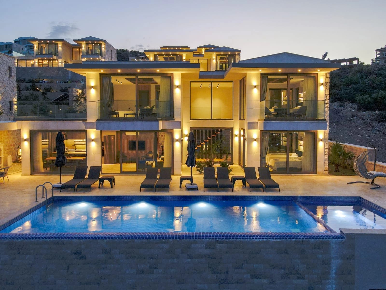 Akşam ışıklandırmalı, önünde dikdörtgen havuz ve şezlonglar bulunan iki katlı modern villa.