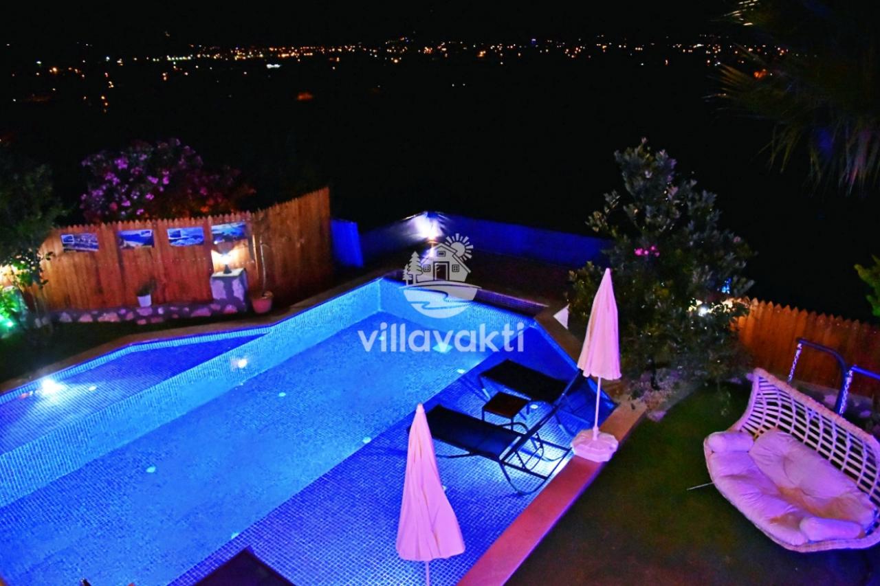 Gece ışıklandırılmış mavi havuz, şezlonglar ve uzaktaki şehir ışıkları manzarası.