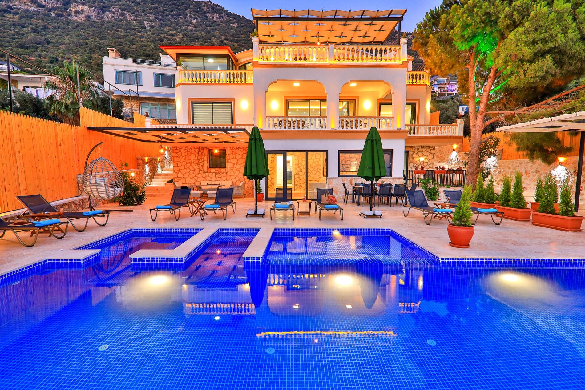 Akşam ışıklandırmalı, havuz başında şezlongları ve yemek masası bulunan iki katlı villa.