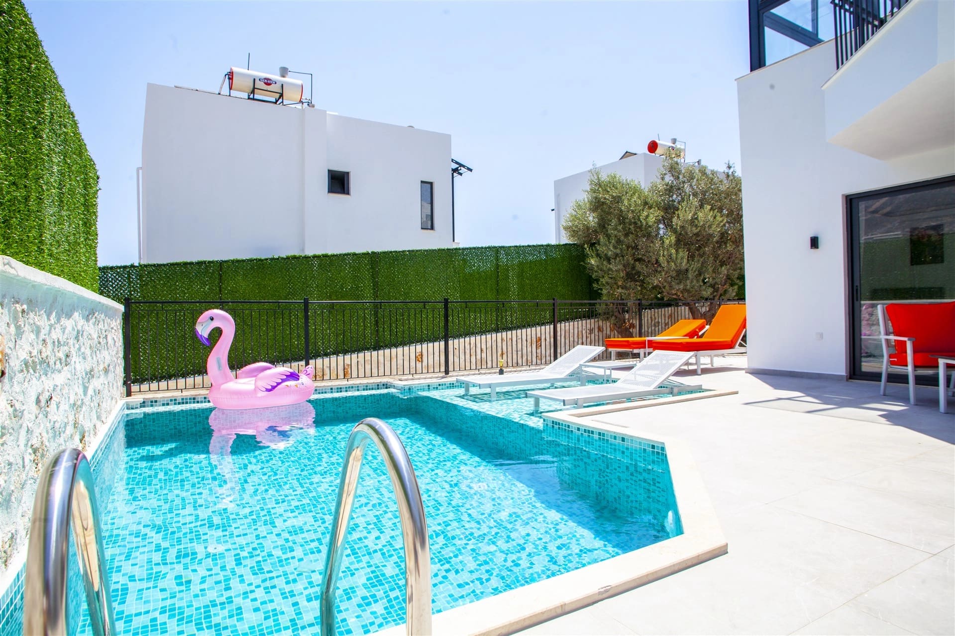 Pembe flamingo deniz yatağı bulunan havuzun kenarında turuncu şezlonglar ve beyaz villa yer alır.