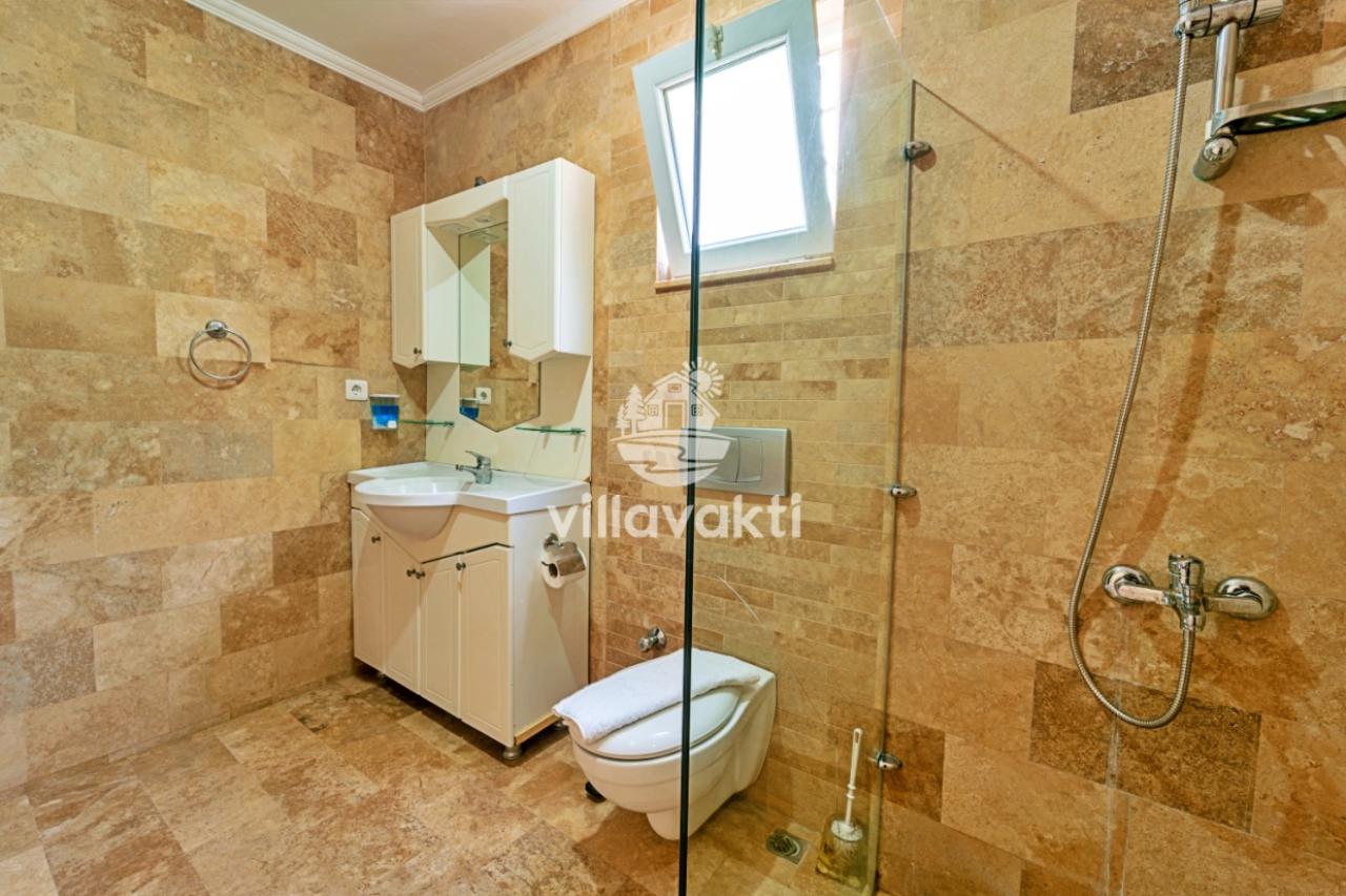 Bej renkli taş kaplamalı banyoda beyaz lavabo, klozet ve cam bölmeli duş alanı bulunur.
