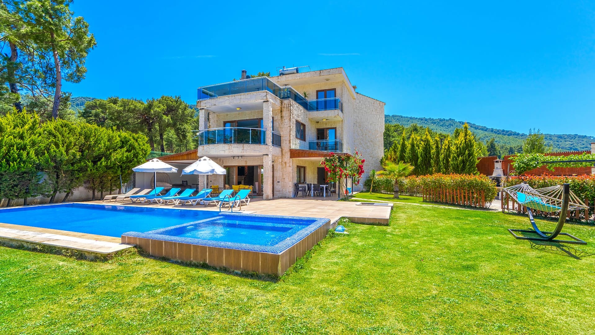 Yeşil ağaçlarla çevrili bahçesinde havuz, şezlonglar ve hamak bulunan üç katlı taş villa.