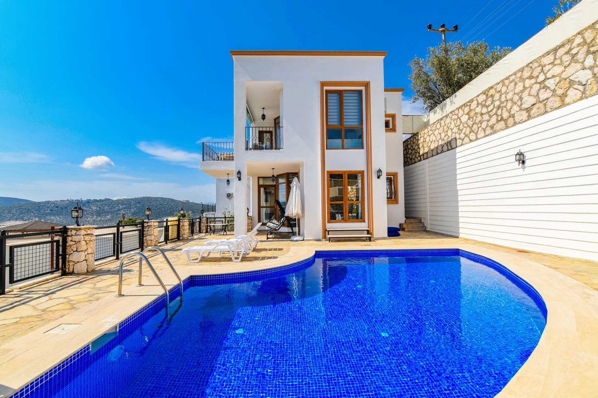 Açık mavi havuzu, şezlongları ve beyaz dış cephesi olan iki katlı villa.