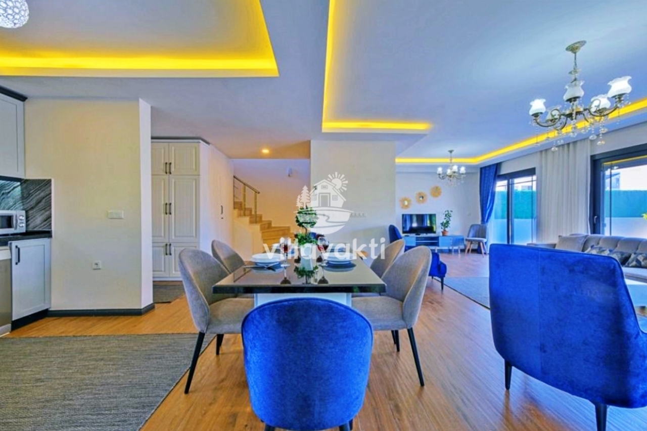 Açık plan salon ve yemek masası, mavi koltuklar ve sarı tavan aydınlatması.