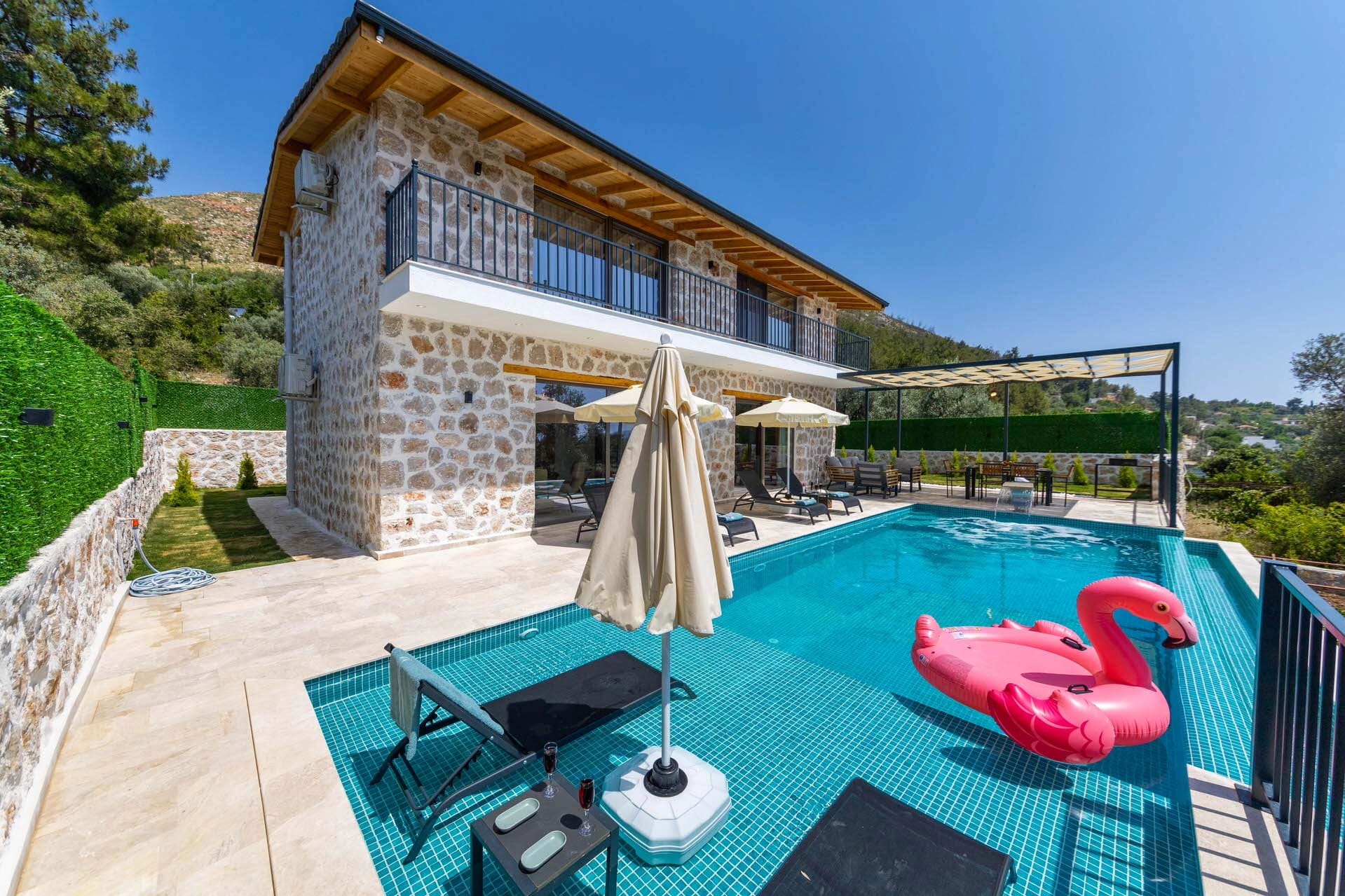 Taş duvarlı iki katlı villa, önünde havuz, pembe flamingo deniz yatağı ve şezlonglar.
