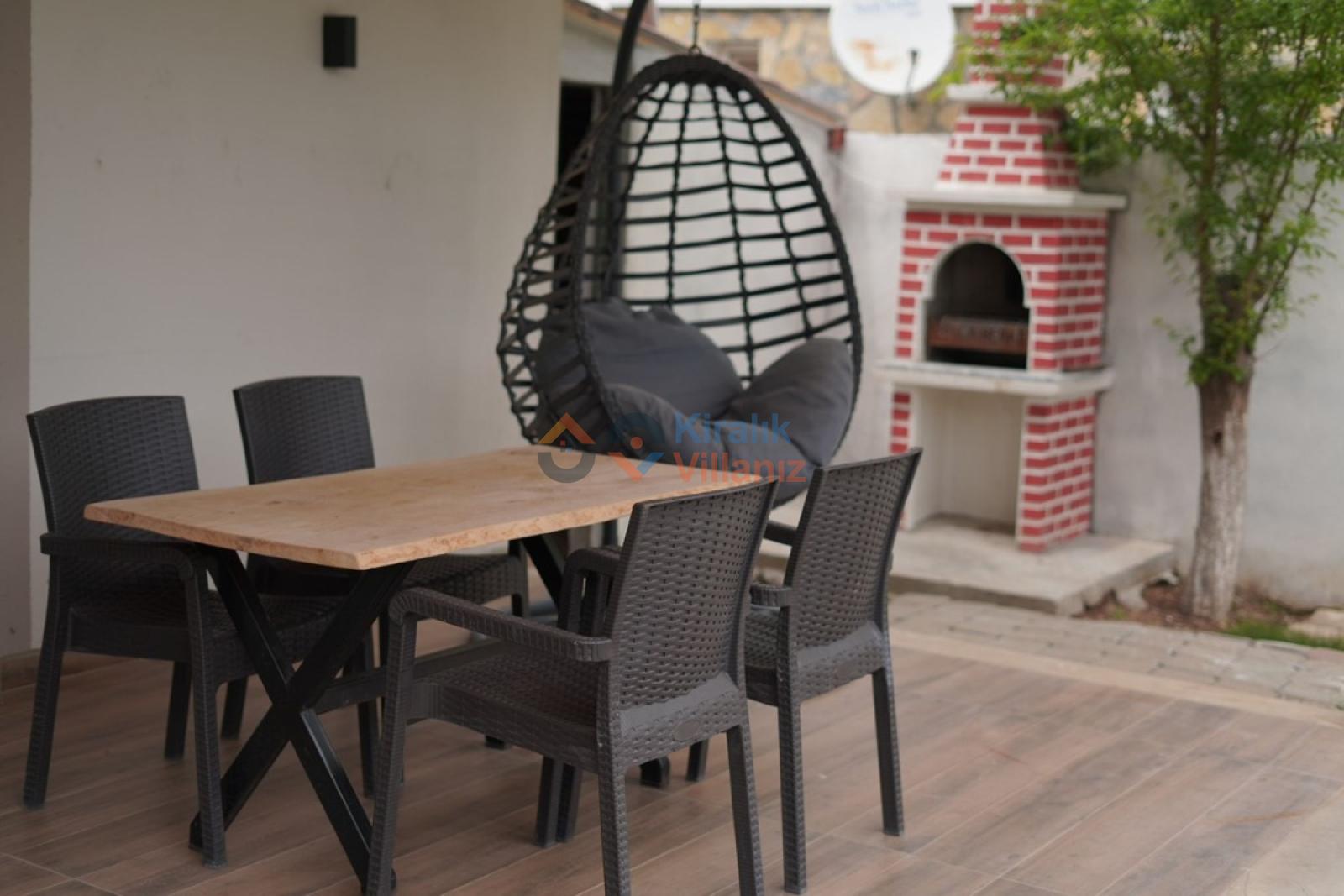 Bahçede ahşap yemek masası, sandalyeler, salıncak ve tuğla barbekü alanı.